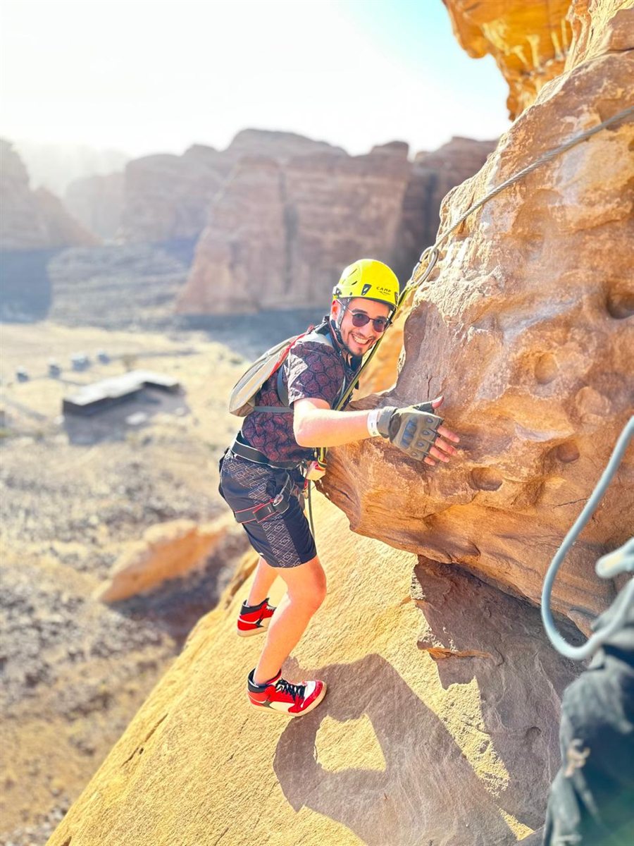 Ilyes in a helmet climbing a sandstone rock face in AlUla