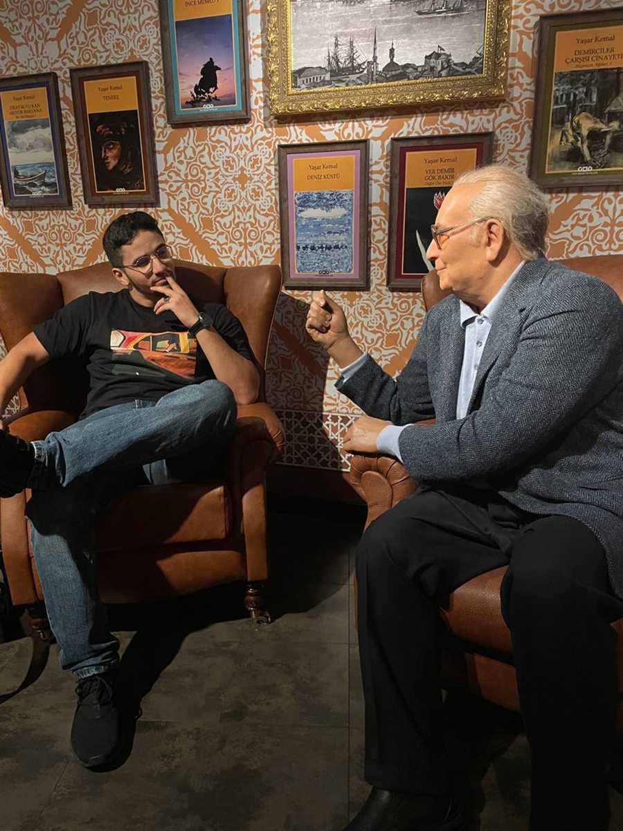 Ilyes in an armchair chatting with a wax figure of the writer Yaşar Kemal